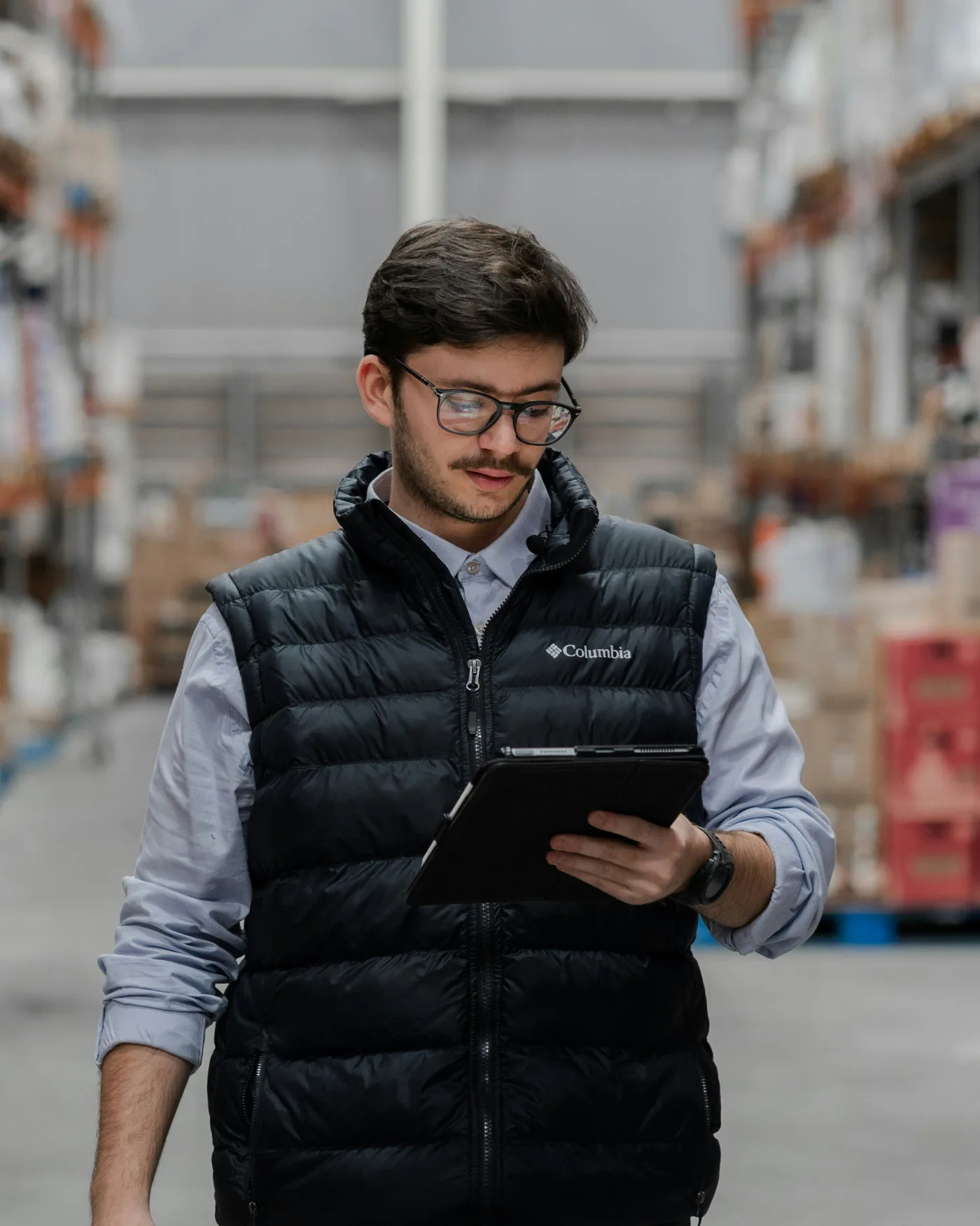 Warehouse supervisor in a work vest walking a racking aisle while reading a tablet